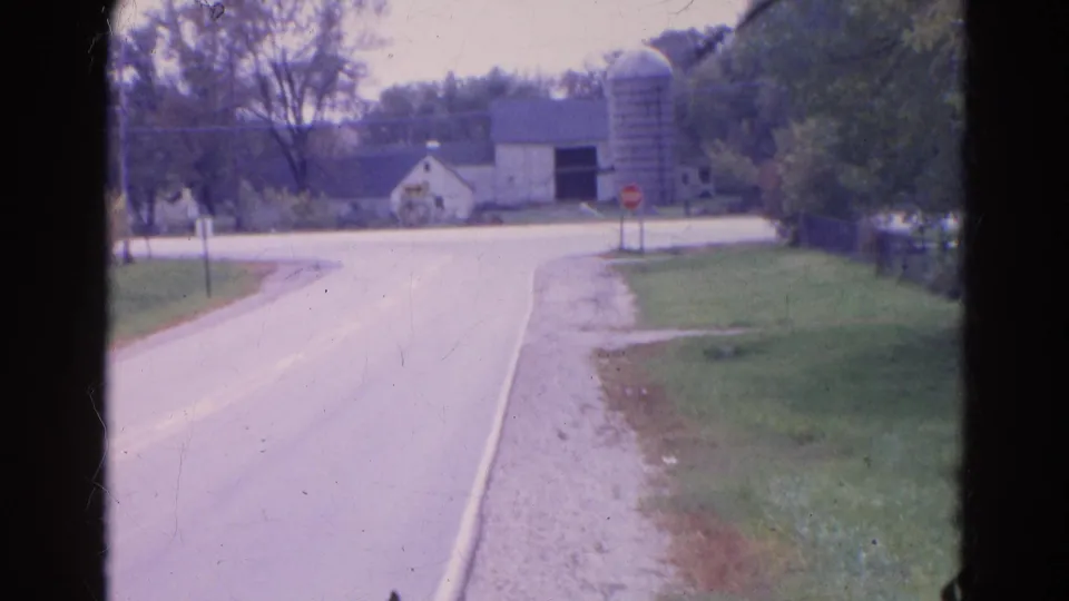 The final frame holds on the Wauconda road approach with a distant car, farmstead, silo, stop sign, trees, and grassy roadside.