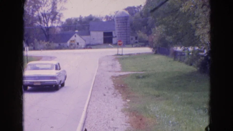 A light-colored sedan passes through the 1968 Wauconda rural road scene, with farm buildings and a stop sign ahead.