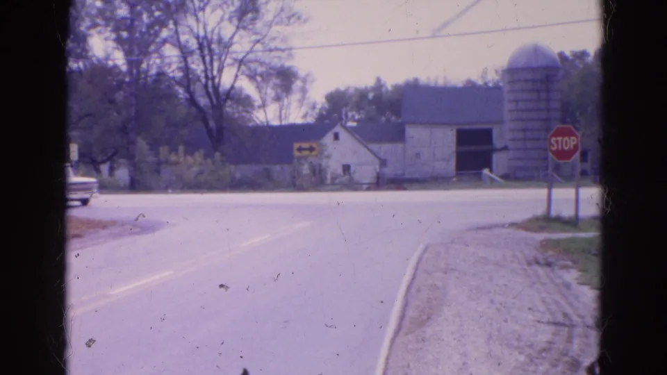 Closer 1968 Wauconda frame of a rural stop sign beside a road, with barn, silo, trees, and a partial vehicle at left.