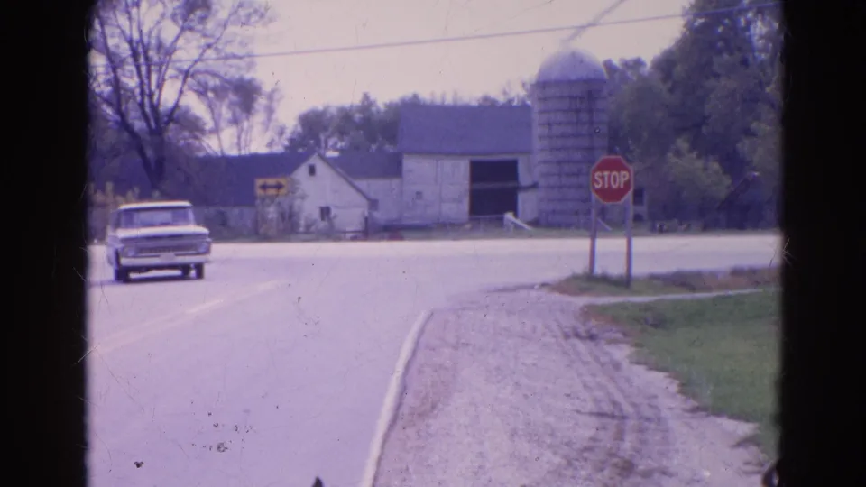 The white pickup continues through the Wauconda rural intersection, with the barn, silo, stop sign, gravel edge, and tree line in view.