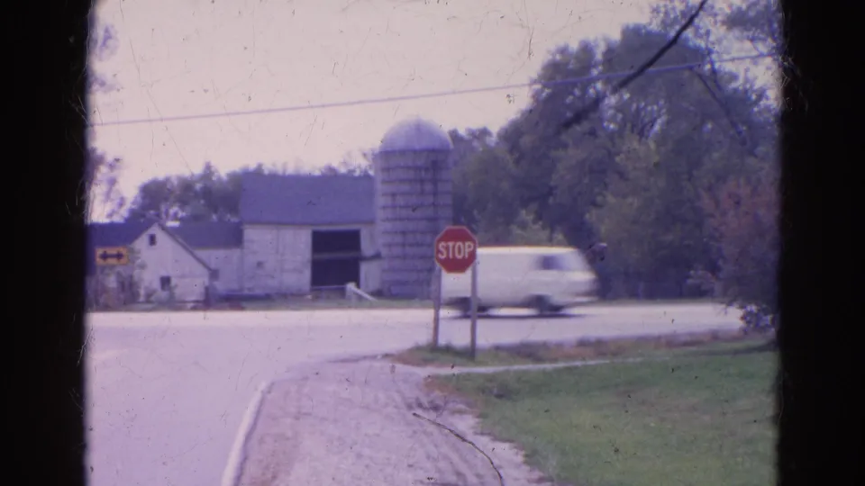 A white pickup passes quickly through the Wauconda intersection, blurring near the red stop sign with the barn, silo, road, and grass verge behind it.