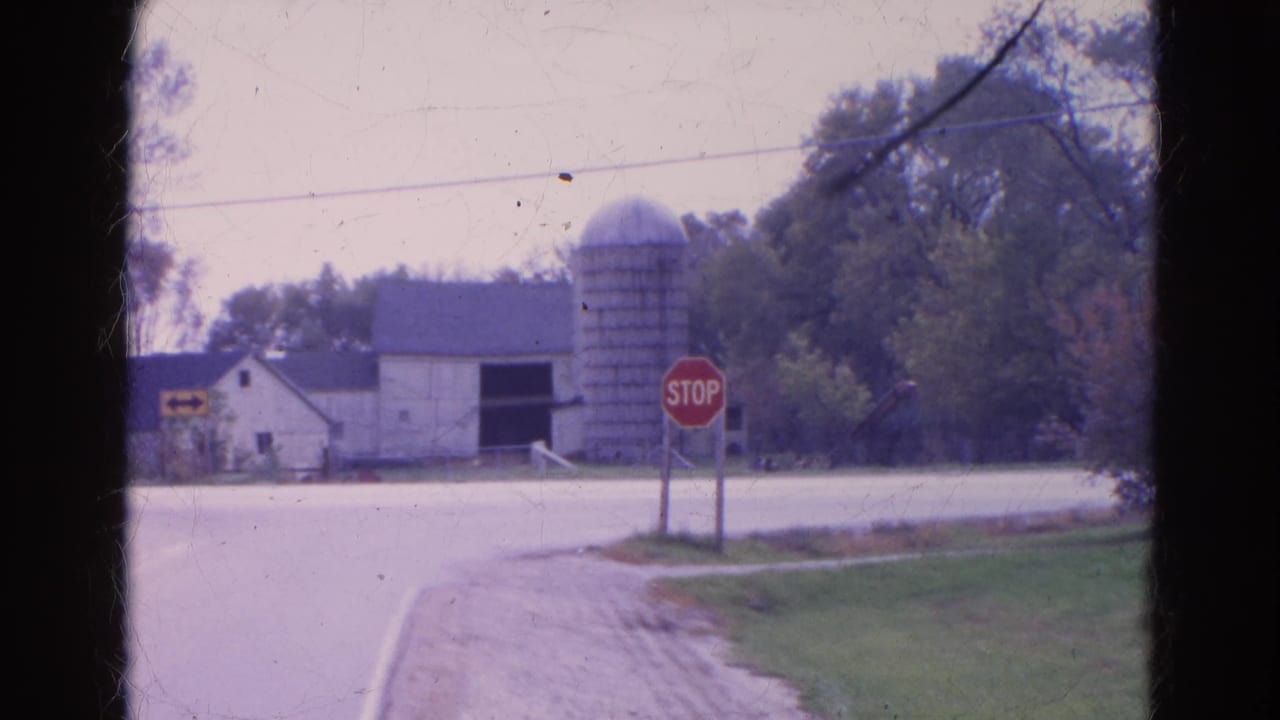 Rural intersection with stop sign, barn, silo, trees, and gravel shoulder.
