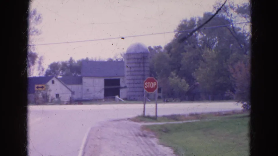 1968 Wauconda, Illinois 8mm frame showing a quiet rural intersection with a stop sign, barn, silo, trees, and road shoulder.