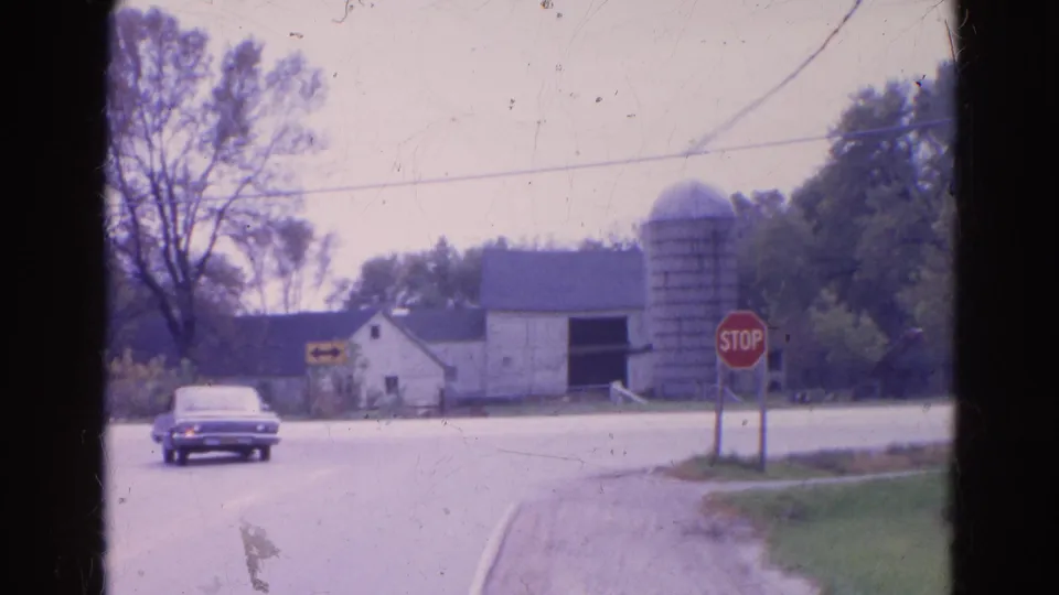 1968 Wauconda, Illinois home-movie frame showing a car entering the rural intersection near a stop sign, barn, and silo.
