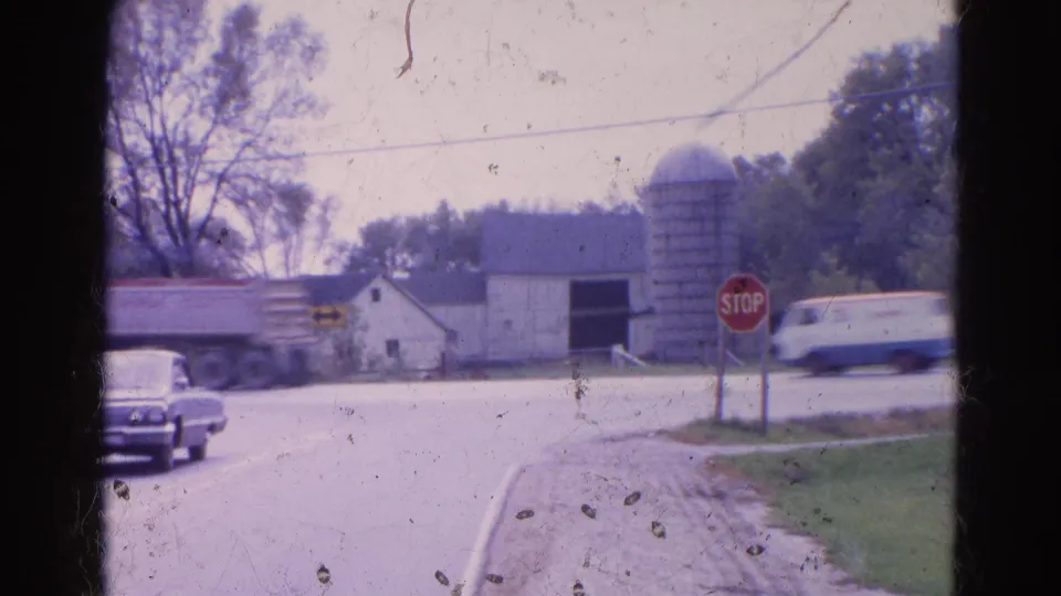 1968 Wauconda, Illinois frame with passing vehicles at a rural stop-sign intersection, with barn, silo, trees, and gravel shoulder visible.