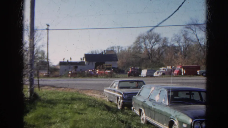 Wauconda, Illinois 1968 home-movie frame showing parked classic cars, roadside grass, overhead wires, and a small building across the street.