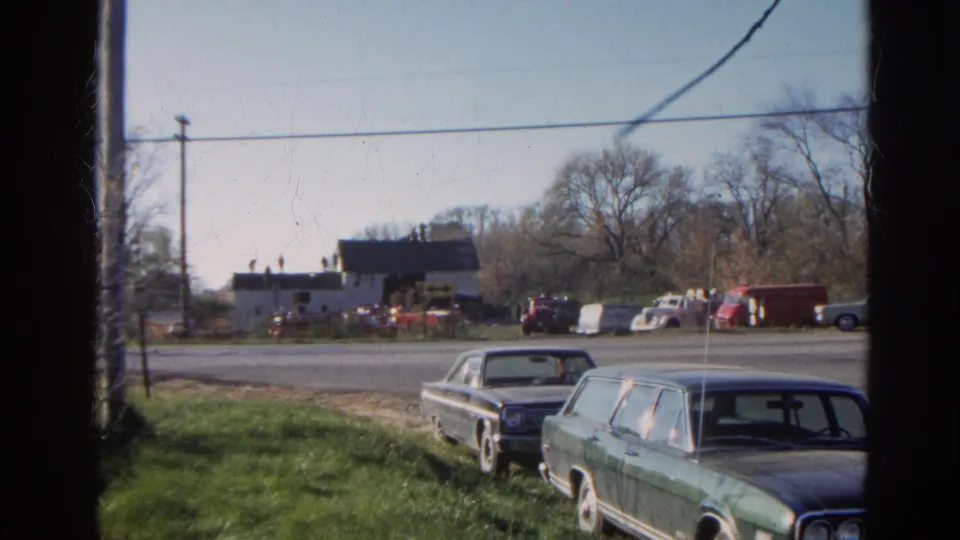 Static 8mm frame from Wauconda, Illinois, showing parked late-1960s cars, a roadside barnhouse, utility lines, and background vehicles.