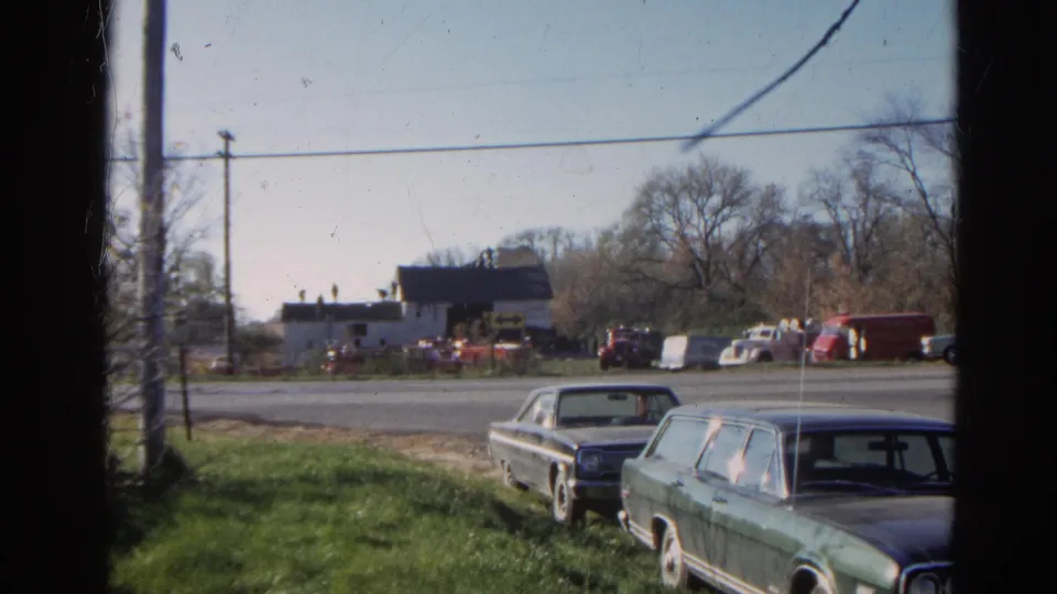 1968 home-movie frame showing a quiet Wauconda roadside with parked classic cars, open grass, utility poles, and a weathered barnhouse.