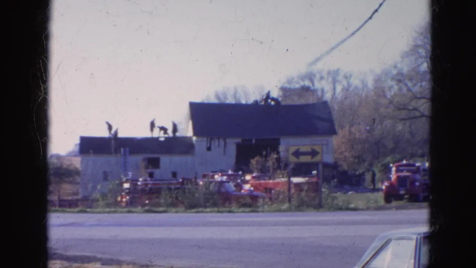 1968 Wauconda, Illinois home-movie frame showing a small white building across a road with figures on the roof and red vehicles nearby.
