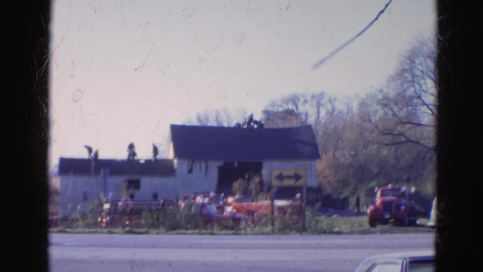 1968 Wauconda, Illinois frame with several distant figures on a dark roof above a white roadside building and red vehicles below.