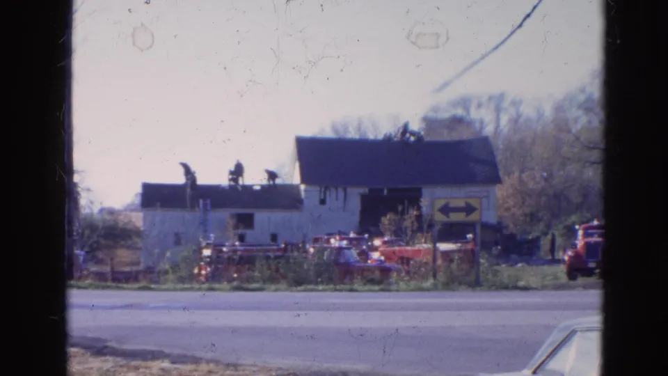 1968 Wauconda, Illinois home-movie view of a roadside building where people appear to work on the roof near parked red vehicles.