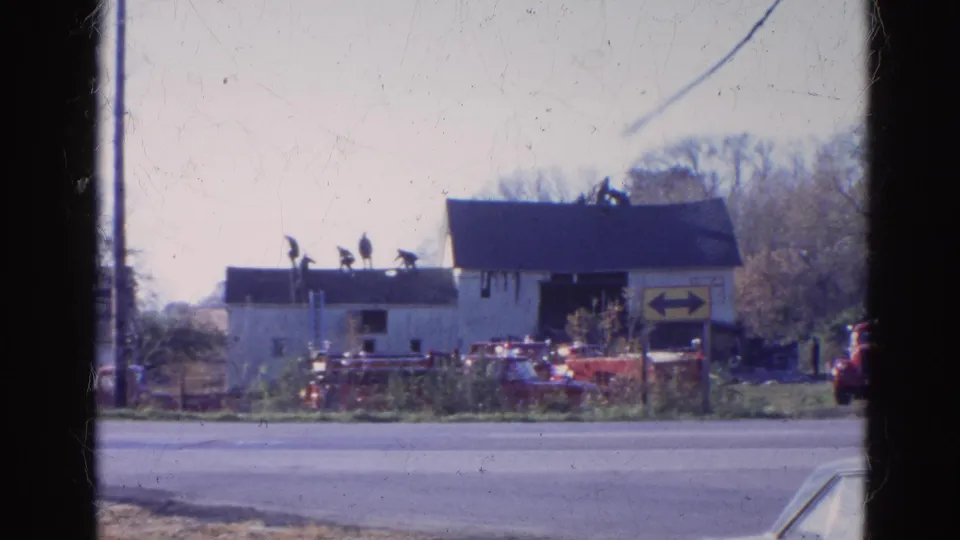 1968 Wauconda, Illinois frame showing multiple distant figures on a roof, red vehicles in the yard, and a yellow directional sign by the road.