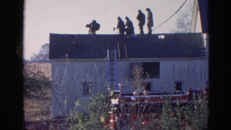 Closer 1968 Wauconda, Illinois frame showing several men on a dark roof, ladders against the building, and red fire equipment below.