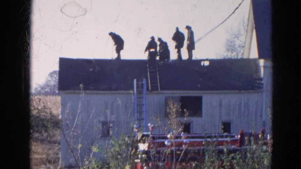 1968 Wauconda, Illinois frame showing men silhouetted on a roof, ladders below them, and emergency equipment in front of the building.
