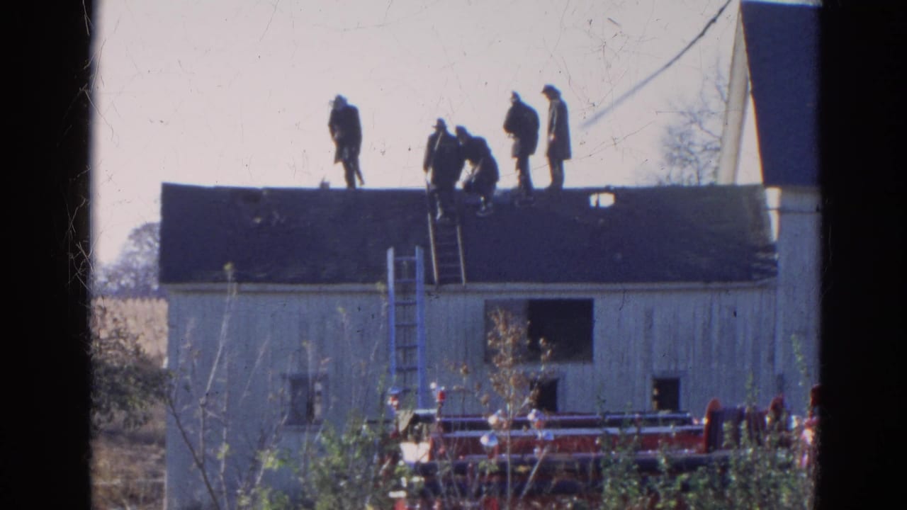 Five men stand and crouch on a roof above a white building with a fire truck below.