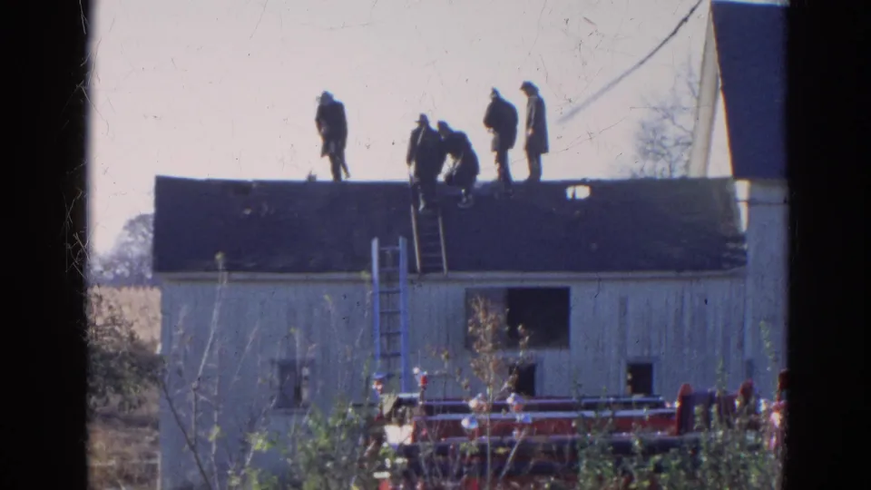 In Wauconda, Illinois, 1968, five men work on the roof of a white wooden building while a red fire truck sits nearby.