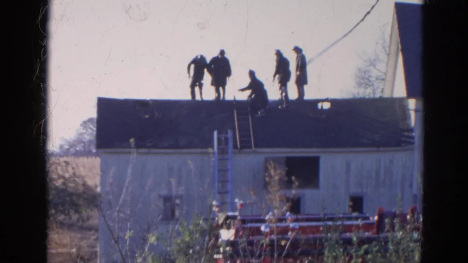 Men continue roof work on the white wooden building in Wauconda, Illinois, with a ladder at center and fire truck equipment below.