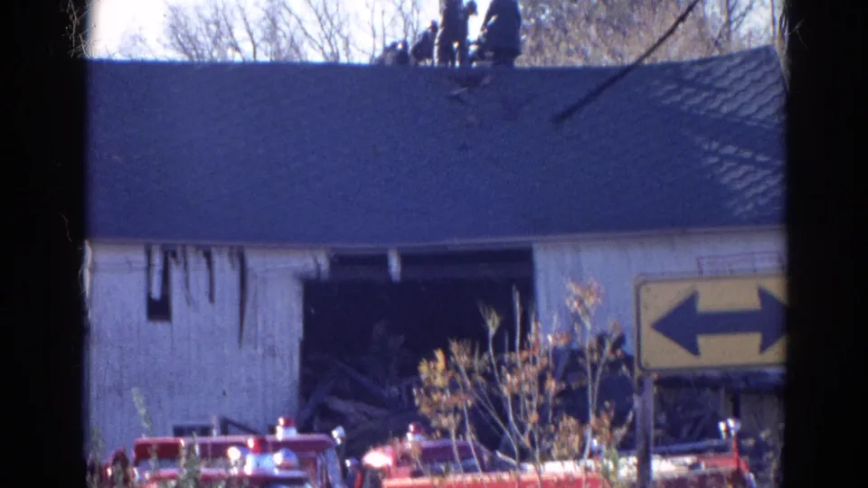 The 8mm frame shows fire trucks below a damaged Wauconda building with firefighters partly visible on the roofline.