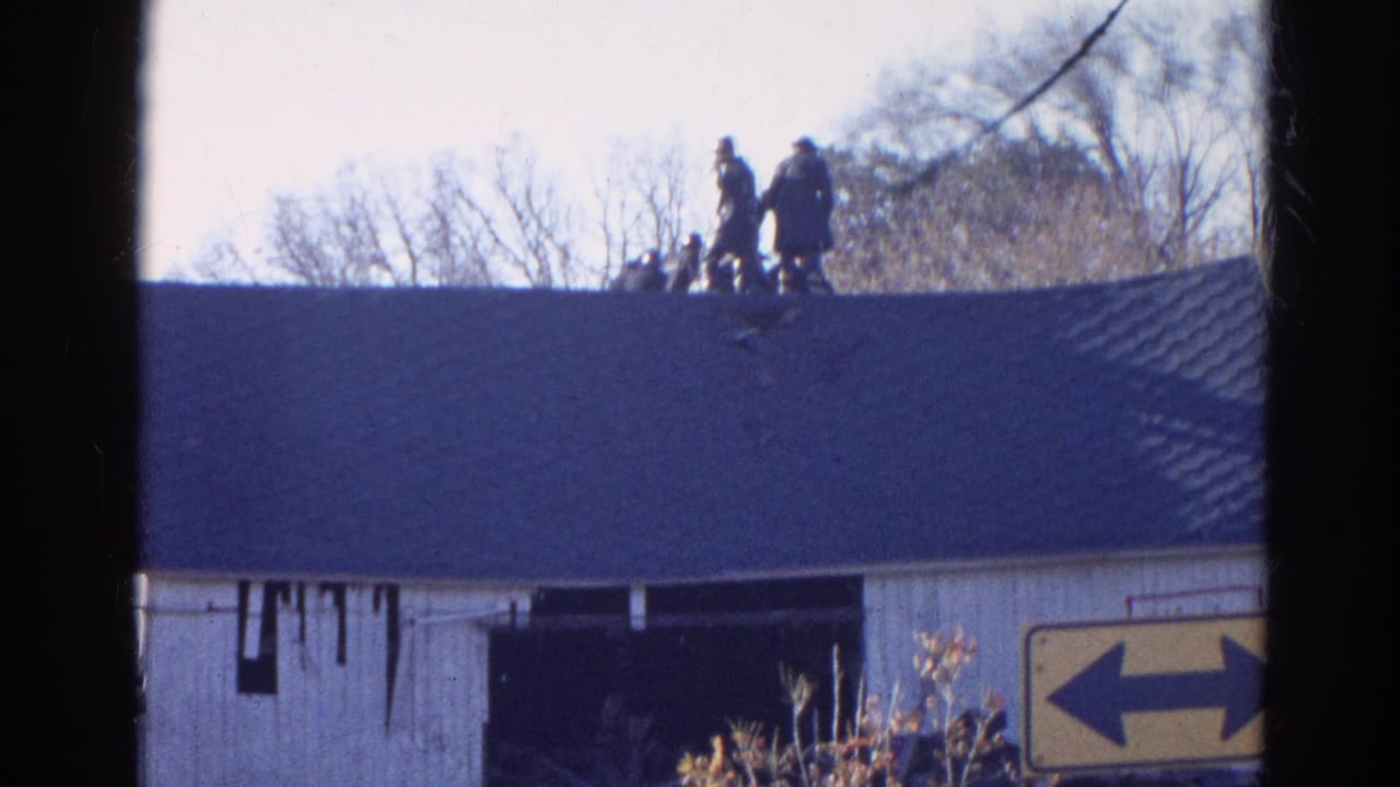 Distant people stand on a dark roof above a white barn wall with bare trees behind them.