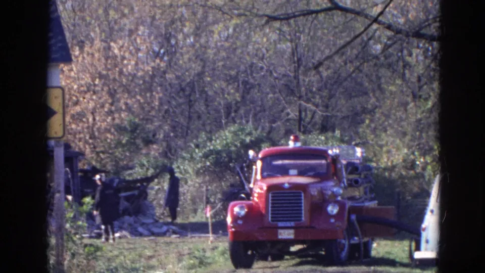 1968 Wauconda, Illinois frame with a red fire truck, wooded background, distant responders, and part of a pale vehicle at right.