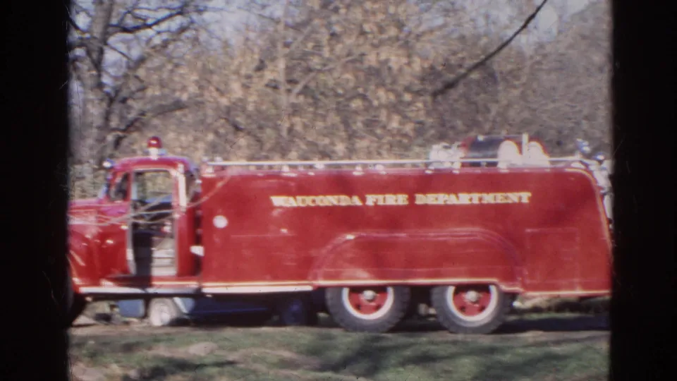 1968 Wauconda, Illinois frame showing the side of a red Wauconda Fire Department truck with open cab door, hose bed, and trees behind it.
