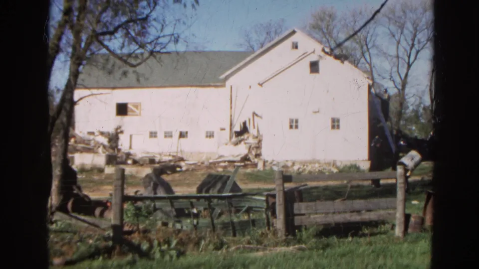 1968 Wauconda, Illinois 8mm frame of a white barn under demolition or renovation, seen past a wooden fence and grassy foreground.