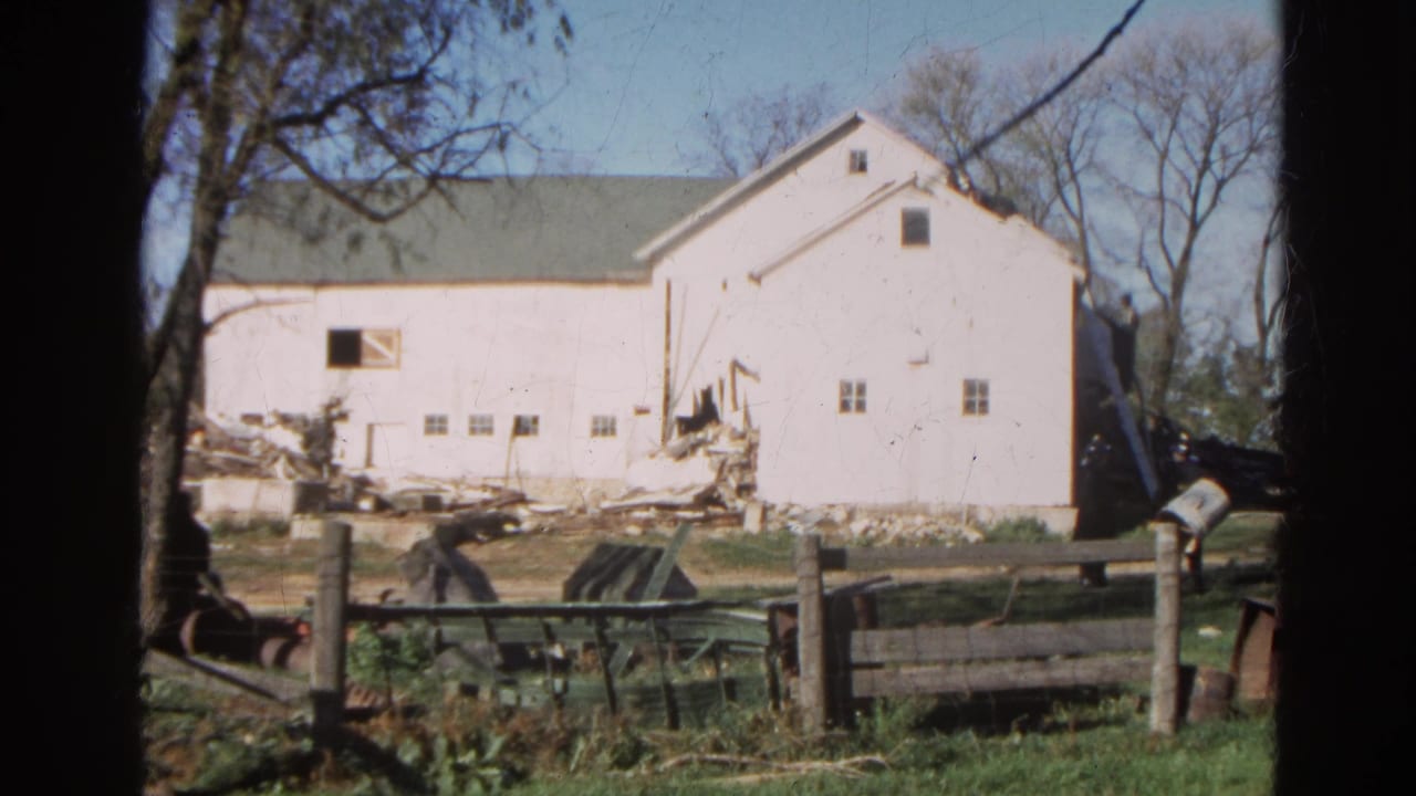 White barn with debris piles, fence posts, grass, and bare trees under a blue sky.