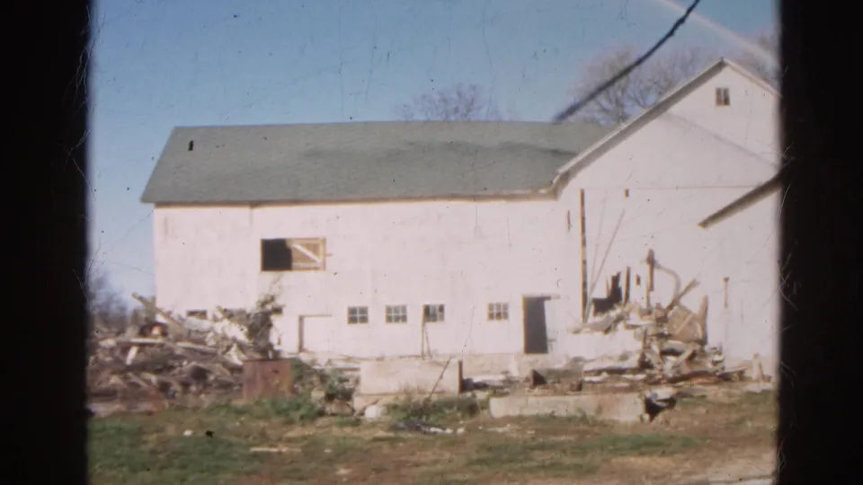 Wide 1968 Wauconda farm frame showing the white barn, scattered demolition debris, open ground, and adjacent roof edge.