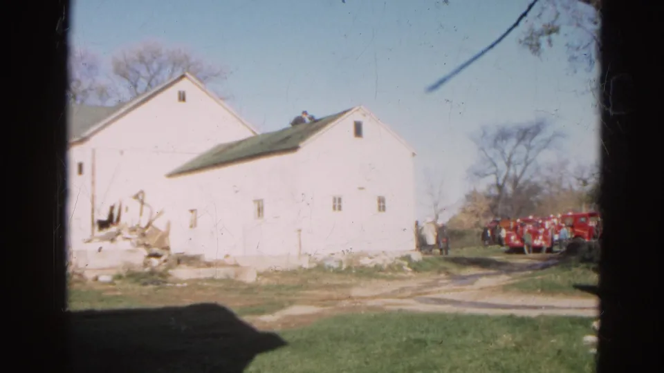 1968 Wauconda, Illinois home-movie frame with debris by a white farm building and fire trucks beyond the driveway.