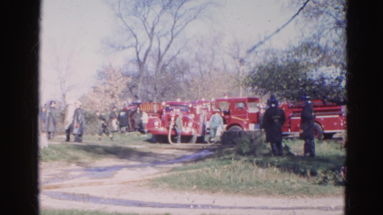 Red fire trucks and firefighters gathered beside a large white building on a rural road.