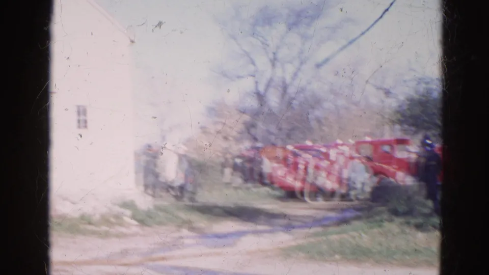 Distant 1968 Wauconda firefighting scene with red trucks lined along a dirt road beside a white building.