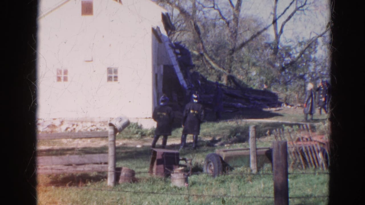 White barn-like building beside firefighters and debris in a grassy outdoor area.