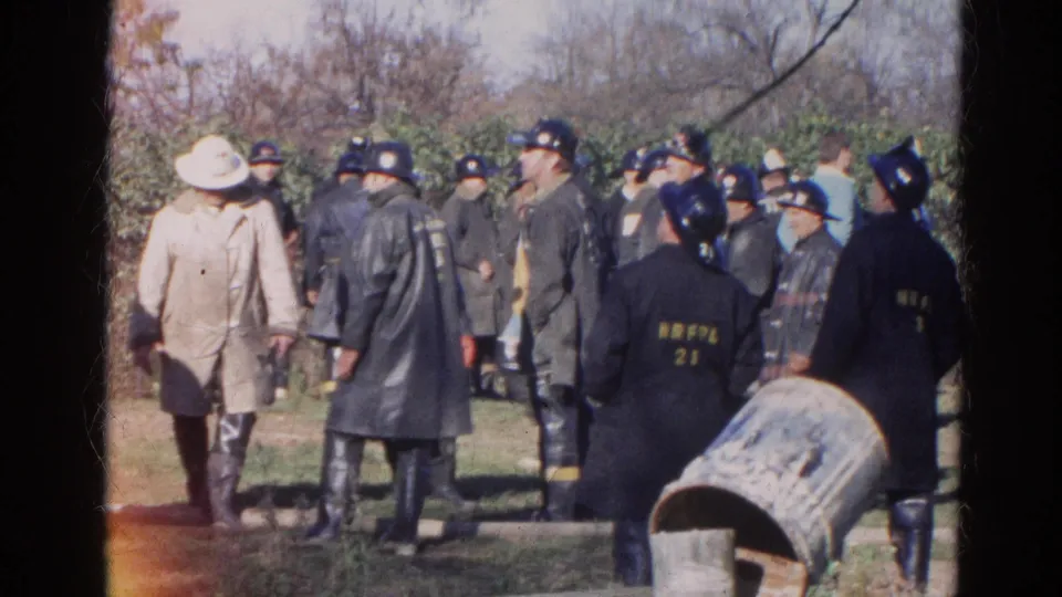 1968 Wauconda frame showing firefighters in dark coats and helmets, a person in a light coat, trees, grass, and an overturned barrel.
