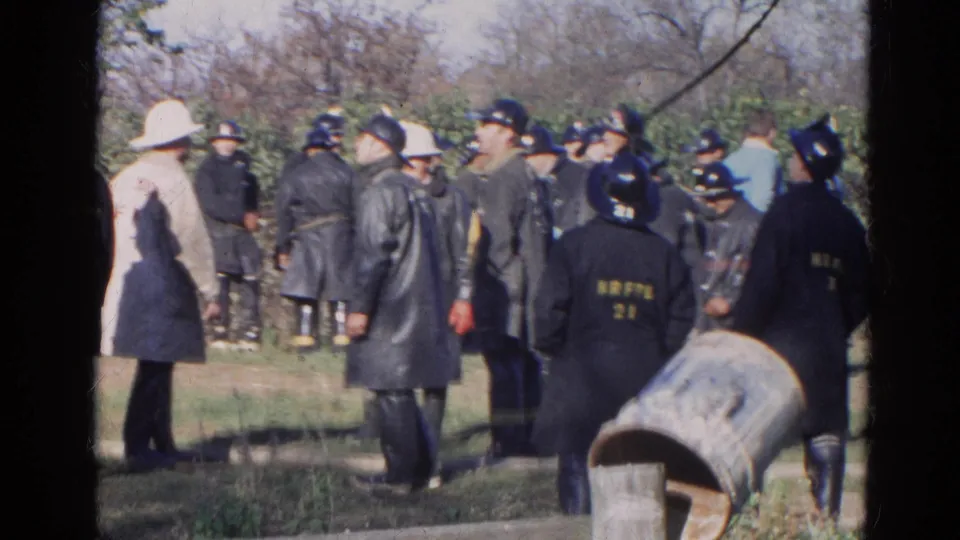 1968 Wauconda 8mm frame with firefighters in dark raincoats, helmet lettering, a light-coated person, bystanders, trees, and grass.