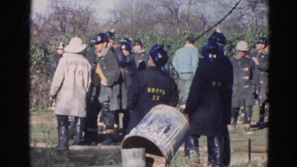 1968 Wauconda footage frame showing firefighters in vintage helmets and coats gathered near a path and trees.