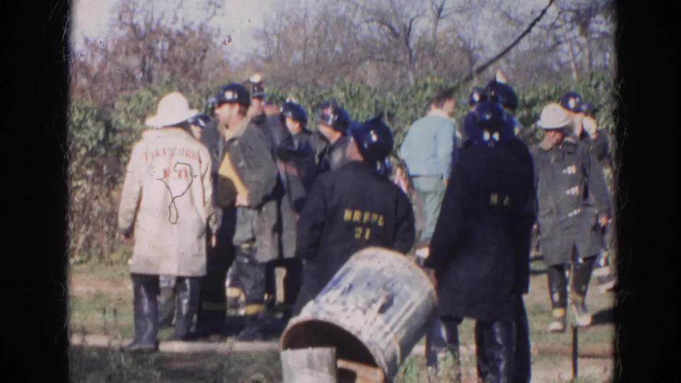 1968 Wauconda frame showing firefighters and uniformed personnel outdoors, with visible coats, helmets, and a metal barrel.