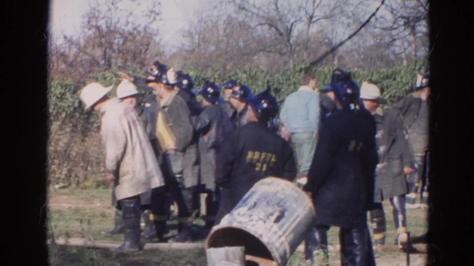 1968 Wauconda, Illinois frame with helmeted emergency workers and a civilian figure gathered outdoors near brush and a barrel.