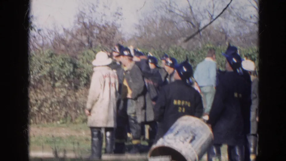 1968 Wauconda, Illinois home-movie frame showing emergency personnel in period coats and helmets huddled near hedges.