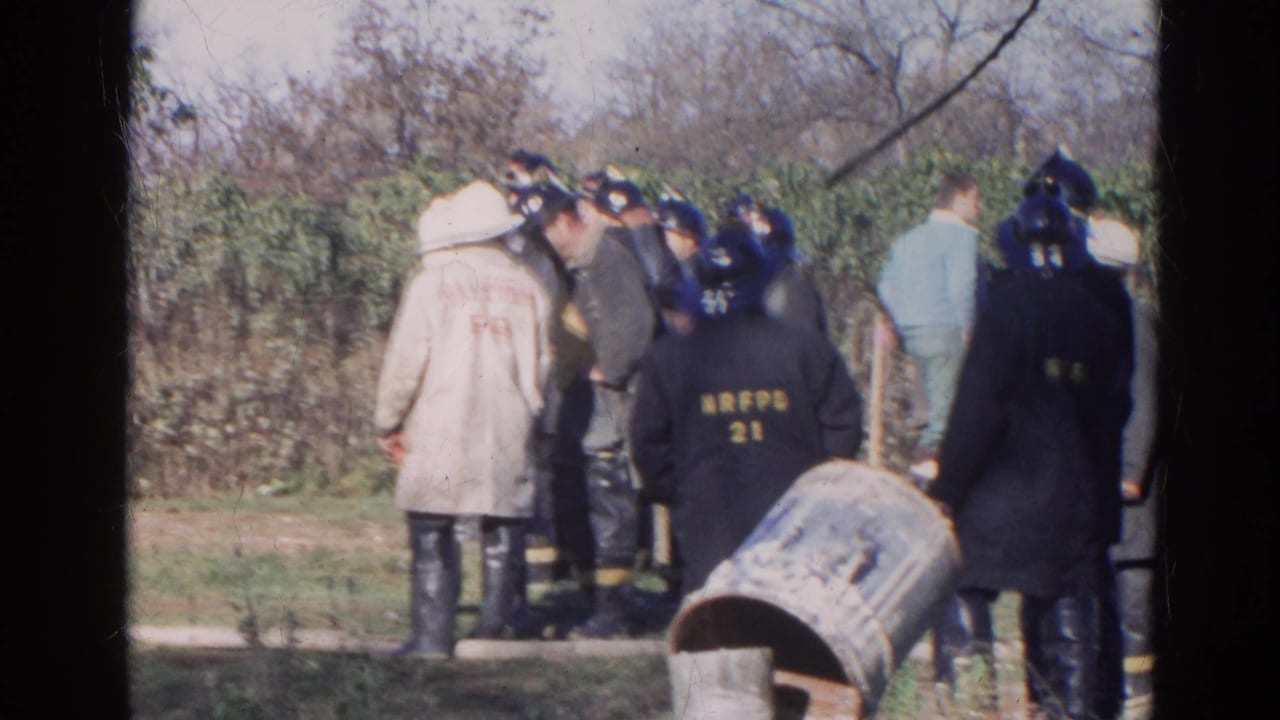 Firefighters and onlookers gather outdoors near brush and a dirt area.
