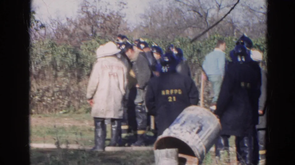 In Wauconda, Illinois, 1968, firefighters in helmets and dark coats stand with onlookers near roadside brush and a metal barrel.