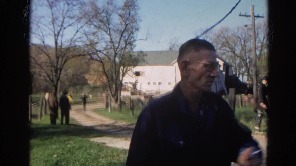 A man in glasses and a dark denim jacket stands before a white barn, dirt drive, trees, and utility pole in Wauconda, Illinois.