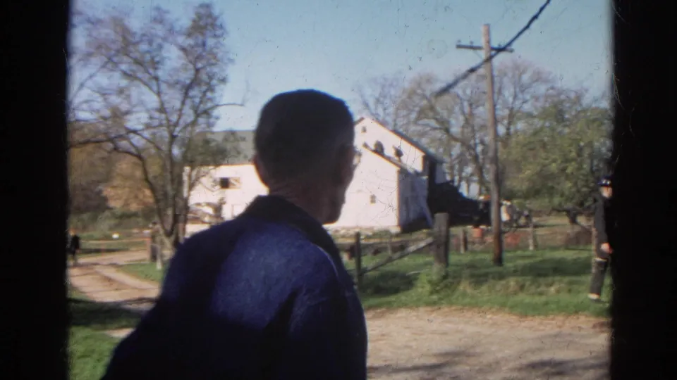 From behind, a man in a dark jacket faces a white barn, dirt drive, utility pole, and small group of people in the rural scene.