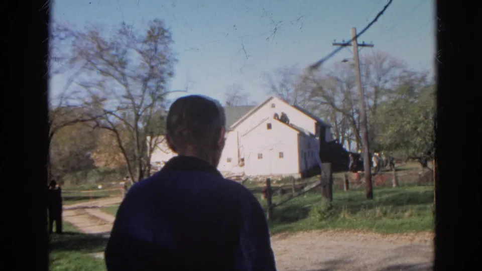 A rear view of the man frames the white barn, dirt path, trees, utility pole, and distant onlookers in the 1968 Wauconda rural setting.