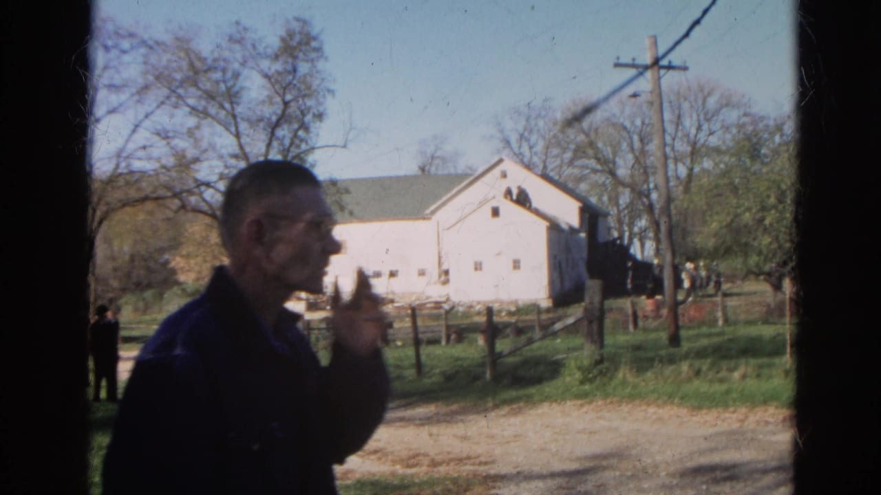 Man in a dark jacket stands near a dirt track with a white farm building behind him.
