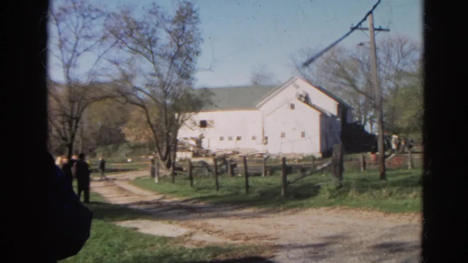 The Wauconda farm setting shows a white building beside a dirt track, with wooden fences, utility poles, trees, and several distant people.