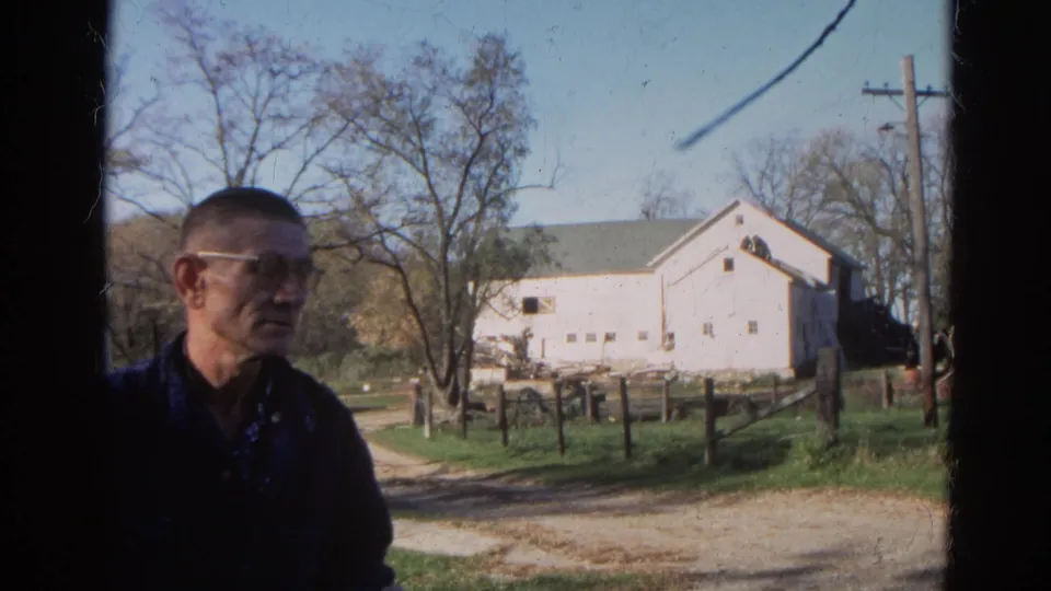 A man in glasses appears in the foreground of the 1968 Wauconda rural scene, with a white farm building, fence line, trees, and track behind him.