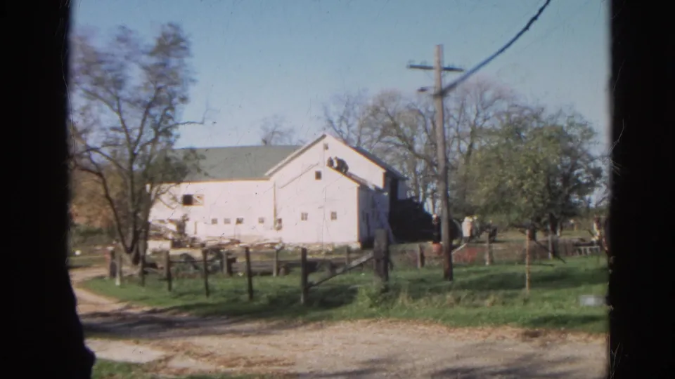 The final frame holds on the Wauconda farm exterior, showing a dirt track, white building, fences, trees, utility pole, grass, and distant figures.