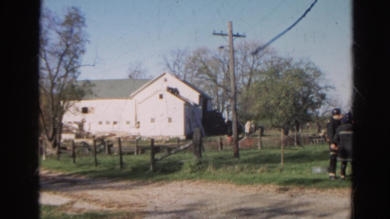 1968 Wauconda, Illinois 8mm frame showing firefighters in dark gear near a white barn, fenced grass, and roadside utility pole.