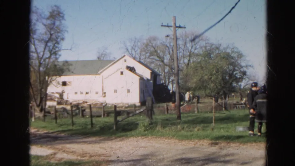 1968 Wauconda, Illinois 8mm frame showing firefighters in dark gear near a white barn, fenced grass, and roadside utility pole.