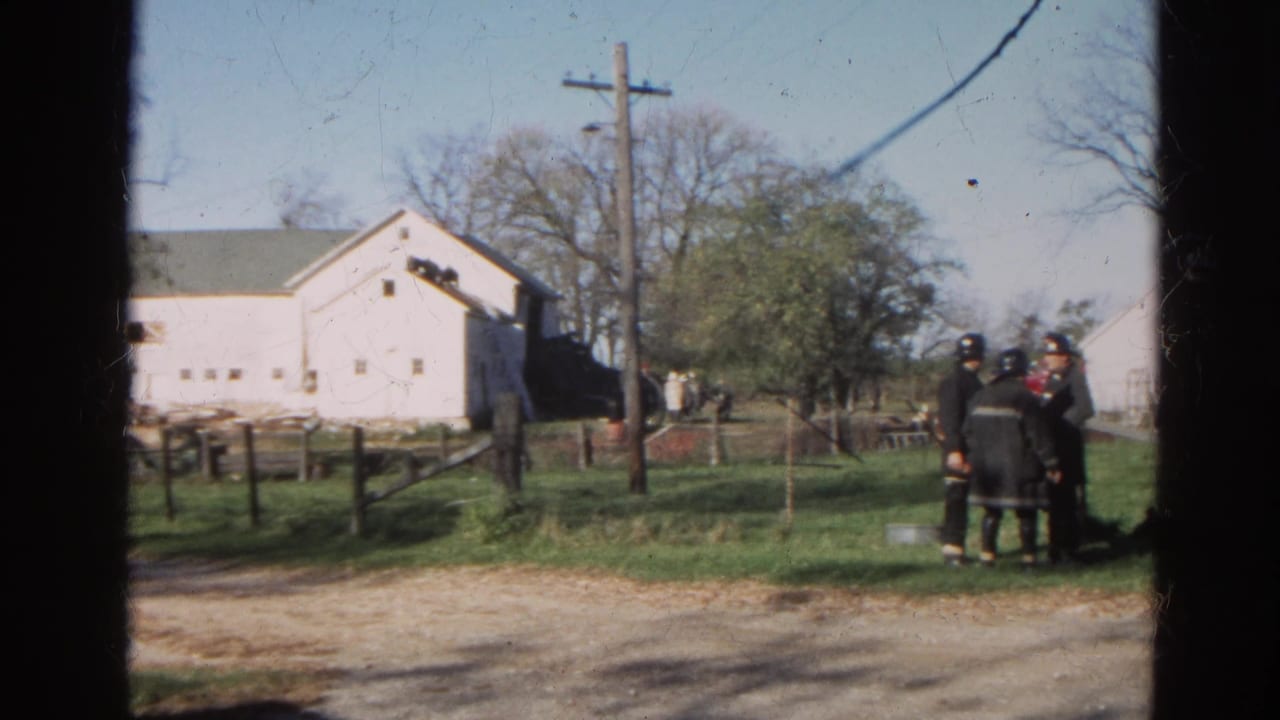 Firefighters in dark turnout gear stand near a grassy roadside with white farm buildings in Wauconda, Illinois, in 1968.